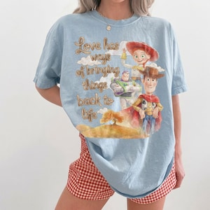 May include: A light blue t-shirt featuring a graphic print of Woody, Buzz Lightyear, and Jessie from Toy Story. The text reads, Love has ways of bringing things back to life, accompanied by a watercolor-style illustration of clouds and a tree.