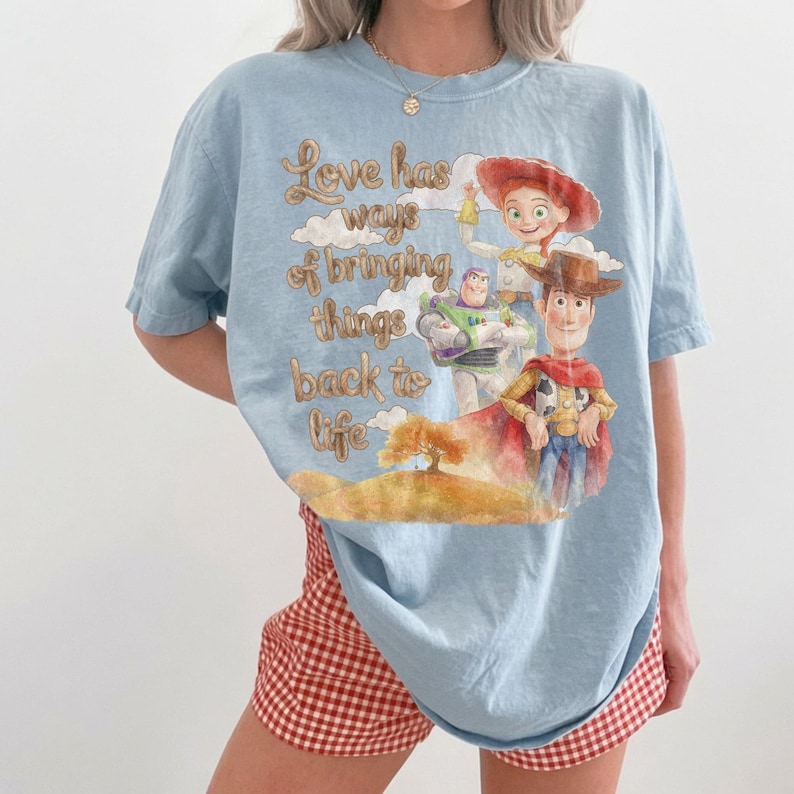 May include: A light blue t-shirt featuring a graphic print of Woody, Buzz Lightyear, and Jessie from Toy Story. The text reads, Love has ways of bringing things back to life, accompanied by a watercolor-style illustration of clouds and a tree.