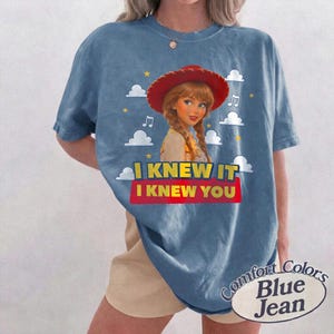 K&ouml;nnte beinhalten: Ein blaues Kurzarm-T-Shirt mit einer Grafik einer Figur mit geflochtenem Haar, die einen roten Cowboyhut tr&auml;gt. Das Design enth&auml;lt wei&szlig;e Wolken, gelbe Sterne, Musiknoten und den Text I KNEW IT I KNEW YOU in Gelb und Rot.
