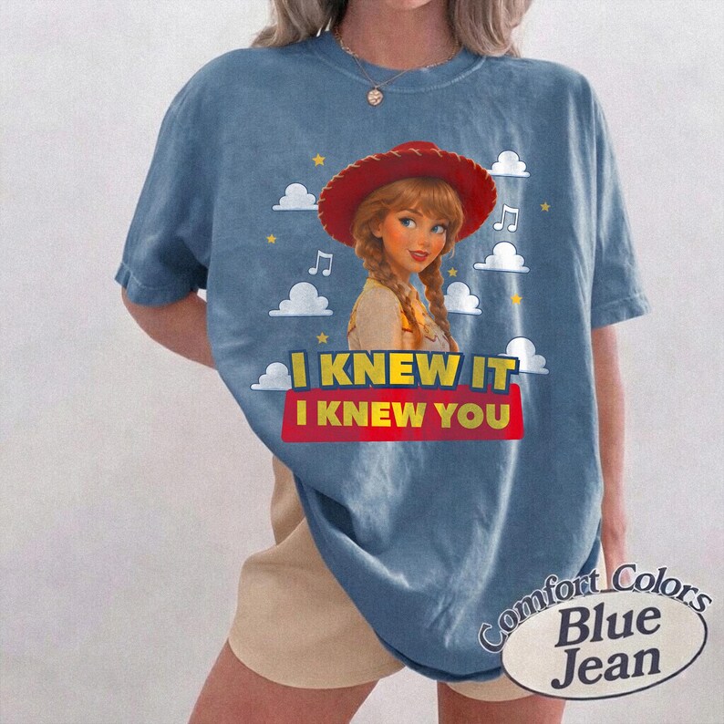 K&ouml;nnte beinhalten: Ein blaues Kurzarm-T-Shirt mit einer Grafik einer Figur mit geflochtenem Haar, die einen roten Cowboyhut tr&auml;gt. Das Design enth&auml;lt wei&szlig;e Wolken, gelbe Sterne, Musiknoten und den Text I KNEW IT I KNEW YOU in Gelb und Rot.