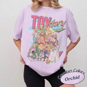 May include: A light purple short-sleeve t-shirt featuring a large graphic print with the text "TOY story" in pink and yellow. The design displays a group of illustrated characters in various outfits, a colorful geometric background, and a ball.