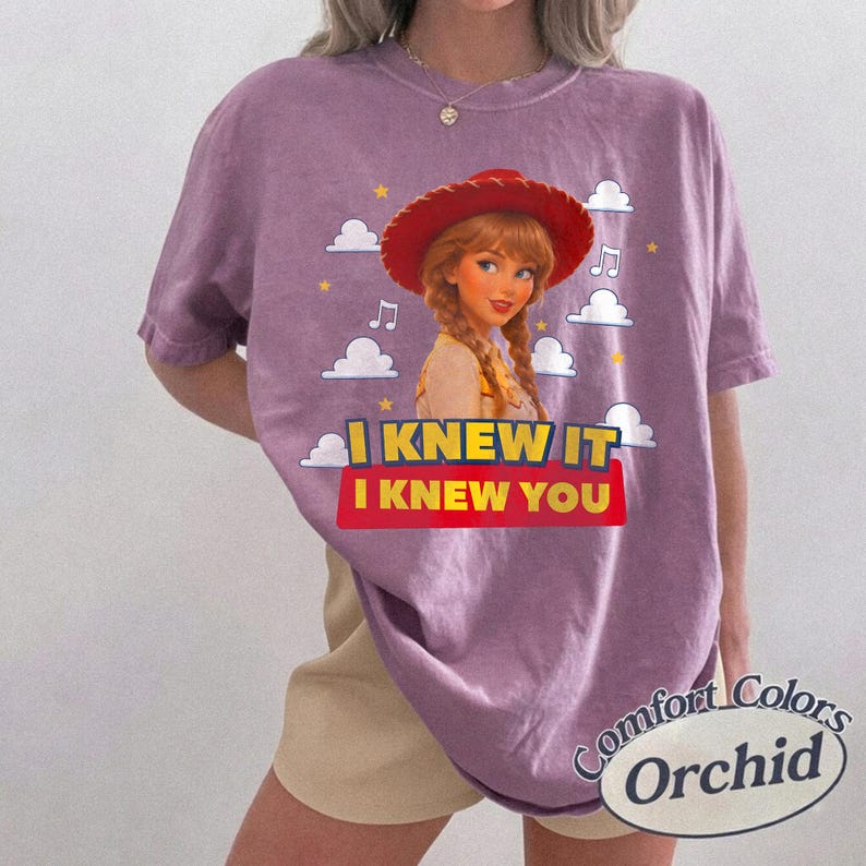 K&ouml;nnte beinhalten: Ein violettes Kurzarm-T-Shirt mit einer Grafik einer Cartoon-Cowgirl-Figur mit geflochtenem Haar und rotem Hut. Das Design zeigt wei&szlig;e Wolken, gelbe Sterne, Noten und den Text I KNEW IT I KNEW YOU in gelben und roten Blockbuchstaben.