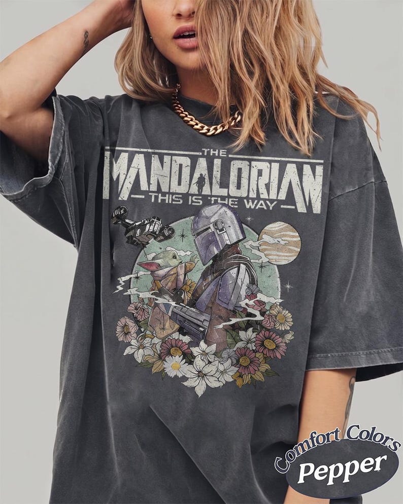 May include: A charcoal gray t-shirt featuring a graphic print of The Mandalorian and Grogu. The text reads THE MANDALORIAN THIS IS THE WAY. The design includes a spaceship, a planet, and colorful flowers. The shirt is labeled Comfort Colors Pepper.