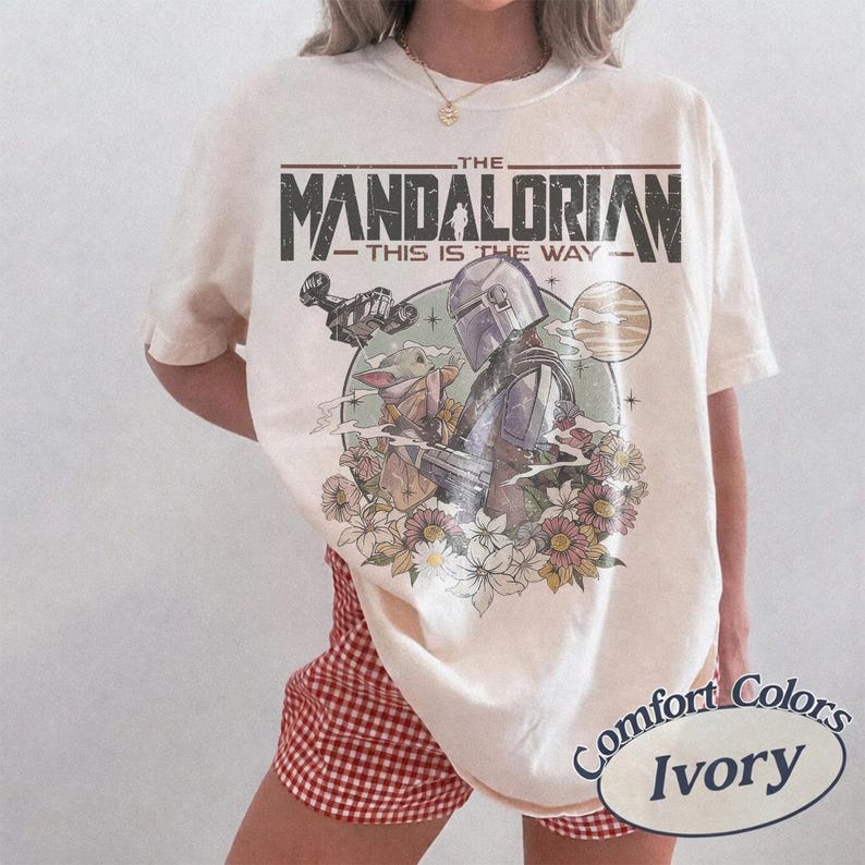 May include: An ivory short-sleeve t-shirt featuring a graphic print of The Mandalorian and Grogu. The design includes the text THE MANDALORIAN THIS IS THE WAY, a spaceship, a planet, and a cluster of flowers in muted tones on the front of the shirt.