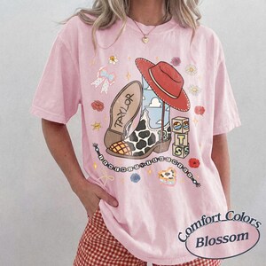 May include: A light pink t-shirt featuring a graphic print of a cowboy boot with a cow print pattern, a red hat, and blocks labeled 13 and TS. The text TAYLOR is on the boot sole, and a beaded string reads I KNEW YOU WERE TROUBLE.