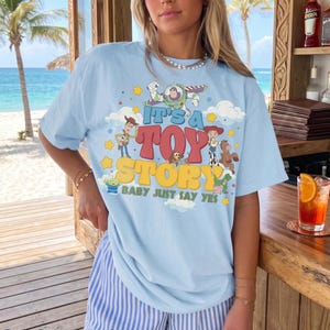 May include: A light blue t-shirt featuring a colorful graphic print of Toy Story characters including Woody, Buzz Lightyear, Jessie, and others. The text reads IT'S A TOY STORY BABY JUST SAY YES in bold, retro-style lettering surrounded by clouds and stars.