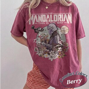 May include: A berry-colored short-sleeve t-shirt featuring a graphic print of The Mandalorian and Grogu. The text reads THE MANDALORIAN THIS IS THE WAY. The design includes a spaceship, a planet, and various flowers in a circular frame on the front.