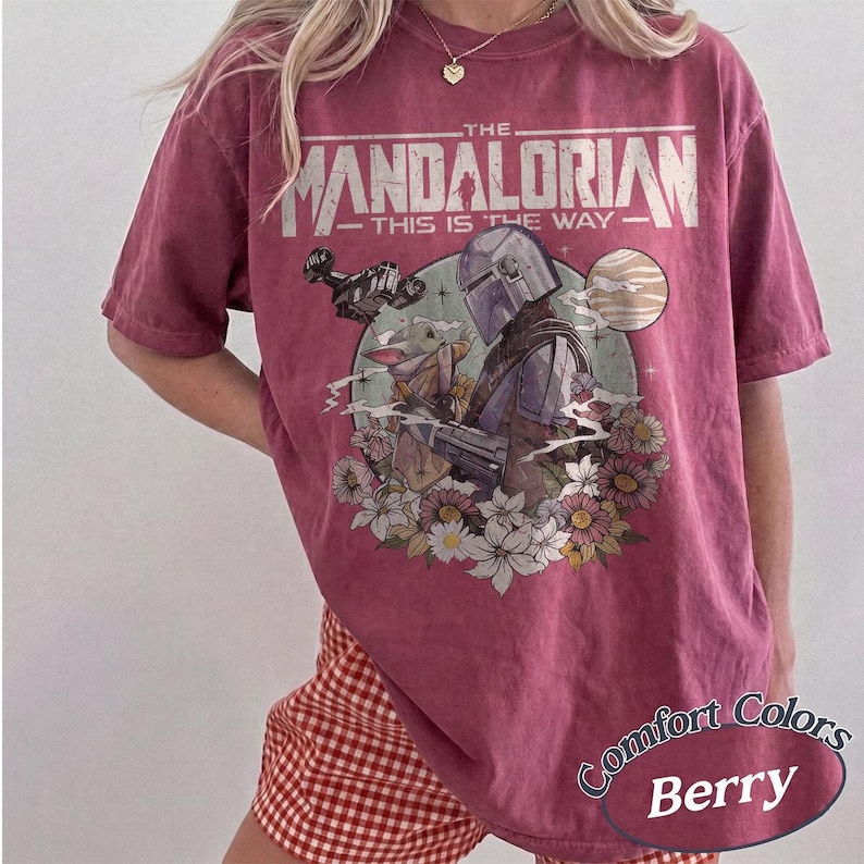 May include: A berry-colored short-sleeve t-shirt featuring a graphic print of The Mandalorian and Grogu. The text reads THE MANDALORIAN THIS IS THE WAY. The design includes a spaceship, a planet, and various flowers in a circular frame on the front.
