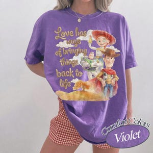May include: A violet short-sleeved t-shirt featuring a graphic print of three animated characters and the text "Love has ways of bringing things back to life". The design includes a cowboy, a space ranger, and a cowgirl set against a landscape.