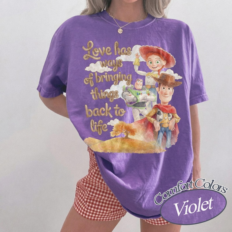 May include: A violet short-sleeved t-shirt featuring a graphic print of three animated characters and the text "Love has ways of bringing things back to life". The design includes a cowboy, a space ranger, and a cowgirl set against a landscape.