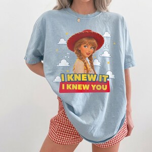 K&ouml;nnte beinhalten: Ein hellblaues T-Shirt mit einer Grafik einer Cowgirl-Figur mit geflochtenem Haar und rotem Hut. Das Design zeigt wei&szlig;e Wolken, gelbe Sterne, Noten und den Text I KNEW IT I KNEW YOU in Gelb und Wei&szlig; auf einem roten Hintergrund.