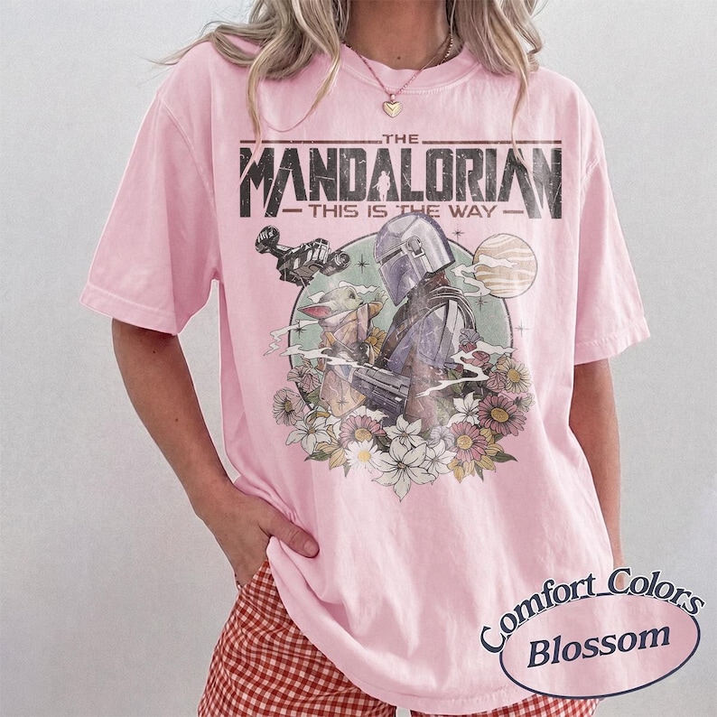 May include: A light pink short-sleeve t-shirt featuring a graphic print of The Mandalorian and Grogu. The text reads THE MANDALORIAN THIS IS THE WAY. The design includes a spaceship, a planet, and various flowers in a circular frame on the chest.