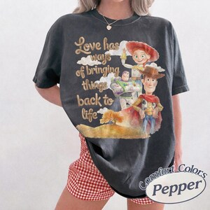 May include: A charcoal gray short-sleeve t-shirt featuring a graphic print of Woody, Buzz Lightyear, and Jessie from Toy Story. The text reads, Love has ways of bringing things back to life. The design includes clouds and a tree on a hill.