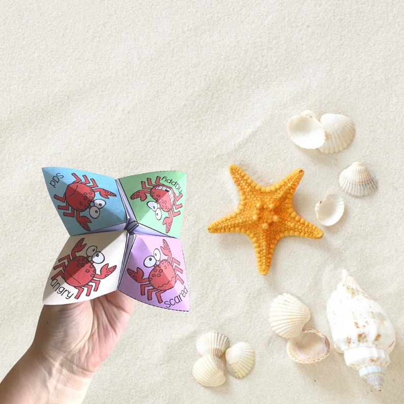 May include: A colorful paper fortune teller with images of crabs and words like "pds", "fiddly", "angry", and "scared". The fortune teller is held in a hand against a background of white sand and seashells.