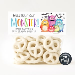 May include: A clear cellophane bag filled with white chocolate-covered pretzels. The bag is sealed with a decorative label featuring colorful cartoon monsters, bats, and stars. Text on the label reads "Build your own MONSTER!" and "Happy Halloween!"