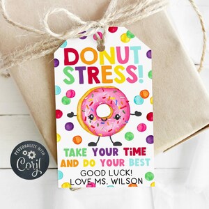 Peut inclure: Une étiquette cadeau avec une illustration de donut rose et les mots "DONUT STRESS!" en couleurs vives. L'étiquette comprend également les phrases "TAKE YOUR TIME AND DO YOUR BEST" et "GOOD LUCK! LOVE MS. WILSON". L'étiquette est attachée à un cadeau emballé avec de la ficelle.