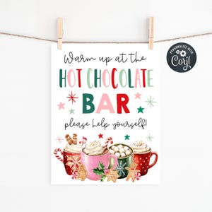 May include: A festive sign with the text "Warm up at the HOT CHOCOLATE BAR, please help yourself!" is displayed. The sign features colorful text and illustrations of hot chocolate mugs with whipped cream and festive toppings. The sign is held by wooden clips.