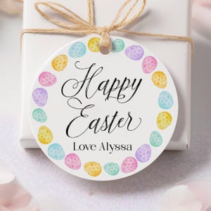 Op de afbeelding: Een witte, ronde paascadeaulabel met de woorden "Happy Easter" in sierlijk schrift, omringd door een rand van pastelkleurige paaseieren. Het label bevat ook de tekst "Love Alyssa". Het label is met touw aan een witte geschenkdoos bevestigd.