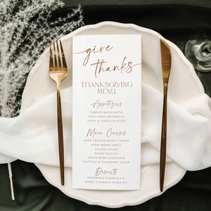 May include: A white rectangular Thanksgiving menu card with copper-colored text reading Give Thanks, Thanksgiving Menu, Appetizers, Main Courses, and Desserts, placed on a white plate between a gold fork and a gold knife on a dark surface.