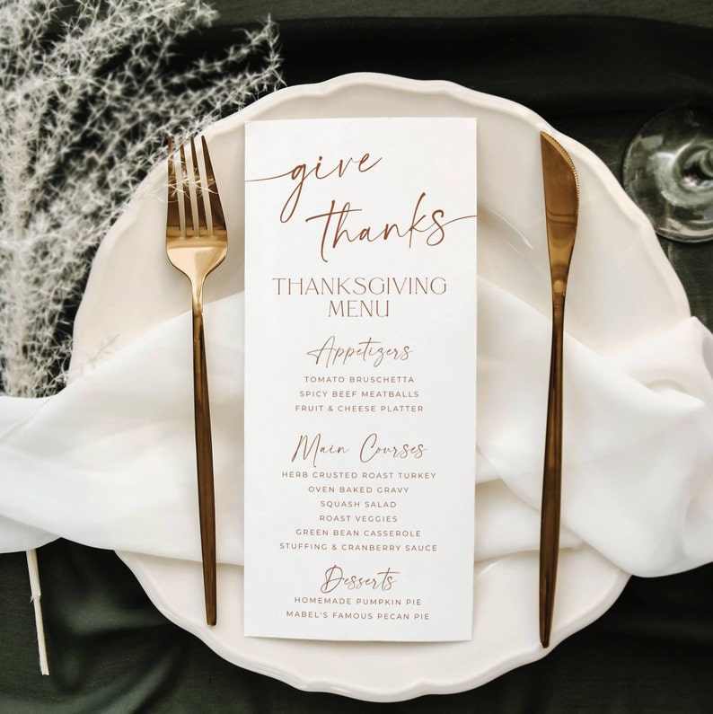 May include: A white rectangular Thanksgiving menu card with copper-colored text reading Give Thanks, Thanksgiving Menu, Appetizers, Main Courses, and Desserts, placed on a white plate between a gold fork and a gold knife on a dark surface.