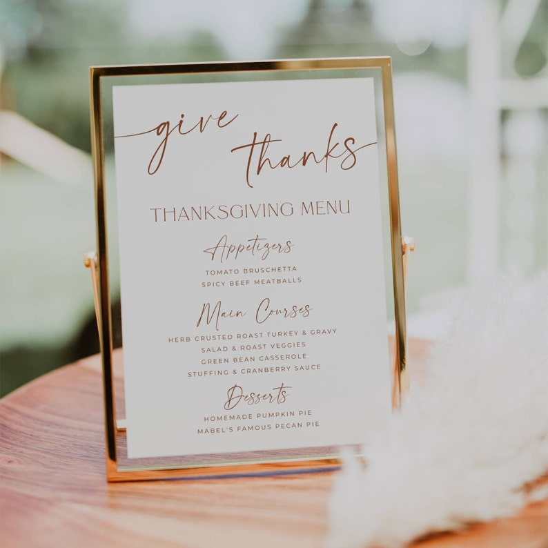 May include: A vertical rectangular menu sign in a thin gold metal frame. The white card features the text "give thanks" in brown script, followed by "THANKSGIVING MENU" and sections for "Appetizers," "Main Courses," and "Desserts" with listed food items.