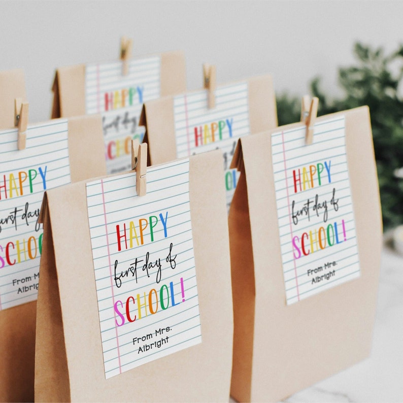 May include: Brown paper bags with colorful "Happy first day of school!" tags attached with clothespins. The tags have a lined paper design and the text is in a rainbow font.