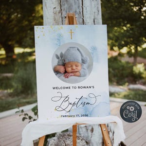 Può includere: Un cartello di benvenuto per un battesimo con uno sfondo ad acquerello in tonalità di blu e oro. Il cartello presenta una foto di un bambino con un cappello grigio e una croce. Il testo sul cartello recita "Benvenuto al battesimo di Rowan il 17 febbraio 2026".