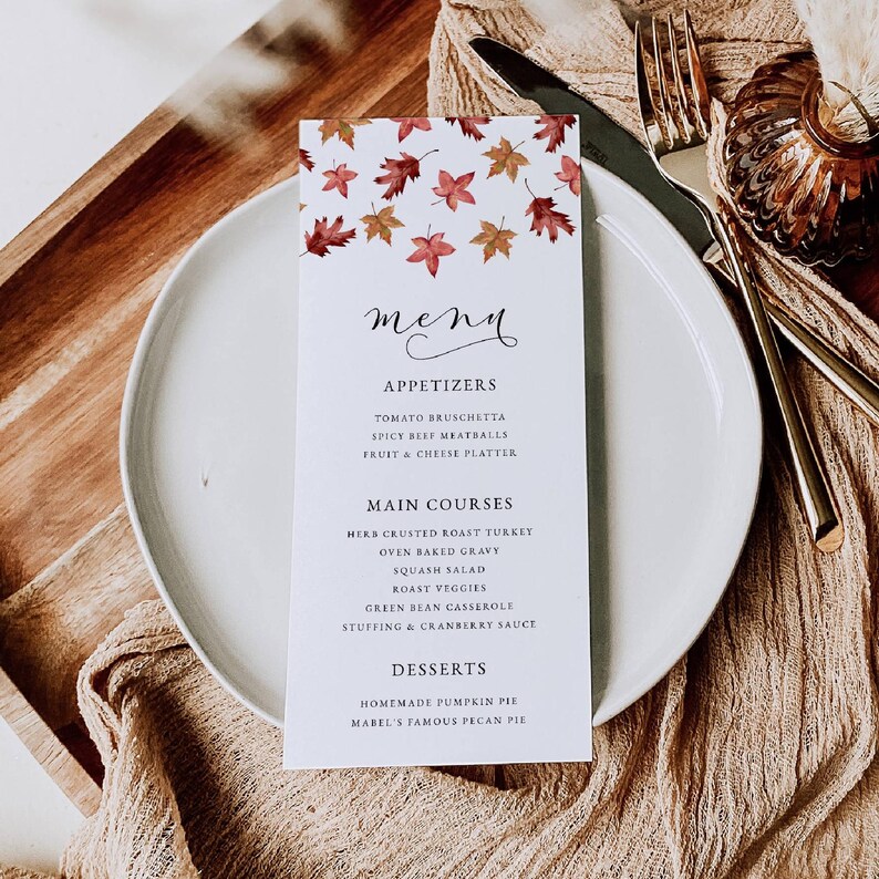 May include: A white menu card with a fall leaf design at the top, featuring appetizers, main courses, and desserts. The menu includes items like herb-crusted roast turkey and homemade pumpkin pie. The card is set on a white plate with silverware.