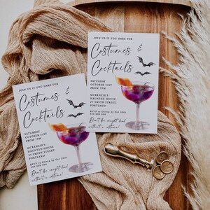 K&ouml;nnte beinhalten: Zwei wei&szlig;e Einladungskarten mit schwarzem Text und einer Aquarell-Illustration eines Cocktailglases mit einem violetten Getr&auml;nk und einer Orangenscheibe. Der Text auf den Karten lautet "Join us if you dare for Costumes & Cocktails" und enth&auml;lt Datum, Uhrzeit, Ort und RSVP-Informationen. Die Karten liegen auf einer Holzoberfl&auml;che mit einem goldenen Siegel und einem goldenen Stoff.