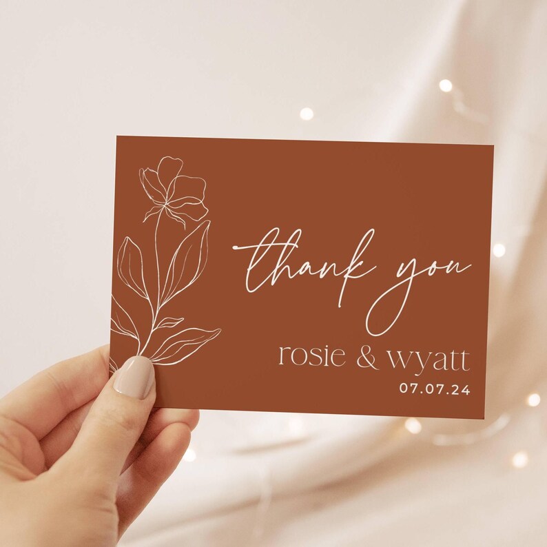 May include: A rectangular thank you card with a terracotta background featuring a white line art floral illustration on the left side. The text reads thank you in script, followed by the names rosie & wyatt and the date 07.07.24 in white.