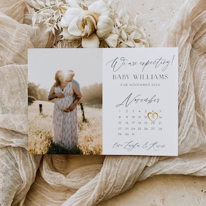 Può includere: Biglietto di annuncio di gravidanza con foto di una coppia in un campo. Il biglietto recita: "We are expecting! BABY WILLIAMS DUE NOVEMBER 2024." Un calendario evidenzia il 13 novembre. Il biglietto è circondato da tessuto e fiori dai toni neutri.