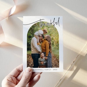 May include: A family of four poses for a photo in a fall setting. The parents are standing with their two children, one on each side. The family is smiling and looking at the camera. The photo is framed in an arch shape with the word "thankful" written above the arch. The text "O'Donnell Family 2022" is written below the arch.
