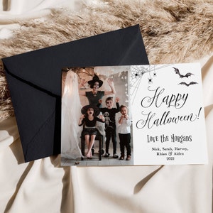 May include: A rectangular Halloween greeting card featuring a photograph of four people in costumes next to a white section with black text that reads Happy Halloween! Love the Morgans, Nick, Sarah, Harvey, Rhian & Aiden 2022, with spiderweb and bat graphics.