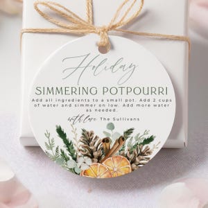 Könnte beinhalten: Ein runder Geschenkanhänger mit dem Text "Holiday SIMMERING POTPOURRI" in grüner Schrift. Der Anhänger enthält Anweisungen und eine botanische Illustration von Tannenzapfen, Zimtstangen und Orangenscheiben. Er ist mit Bindfaden an einer weißen Geschenkbox befestigt.