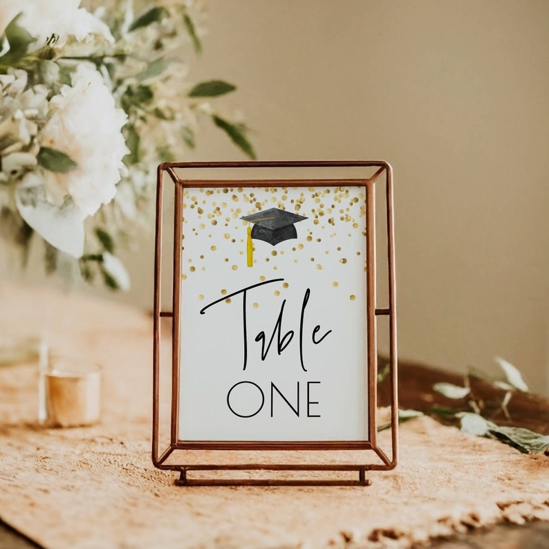 K&ouml;nnte beinhalten: Ein goldfarbener Tischnummernschild mit einer schwarzen Abschluss-Kappe und goldenen Konfetti auf wei&szlig;em Hintergrund. Das Schild zeigt "Table One".