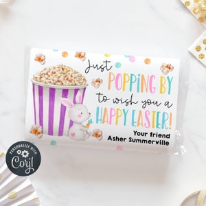 Può includere: Un biglietto bianco con un'illustrazione colorata di un coniglio che tiene una carota accanto a un secchio di popcorn. Il testo dice "Just Popping By to wish you a Happy Easter! Your friend Asher Summerville".