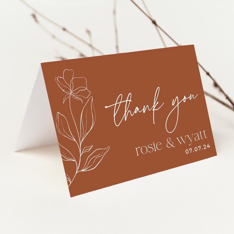 May include: A folded thank you card with a burnt orange background featuring a white line art floral illustration on the left side. The text reads thank you in a script font, followed by the names rosie & wyatt and the date 07.07.24.