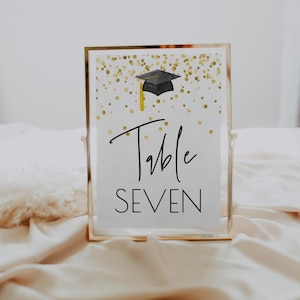 K&ouml;nnte beinhalten: Ein wei&szlig;es Tischnummernschild mit goldenen Konfetti und einer schwarzen Graduation Cap. Das Schild lautet "Tisch Sieben".