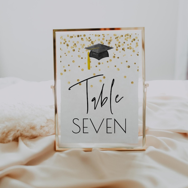 K&ouml;nnte beinhalten: Ein wei&szlig;es Tischnummernschild mit goldenen Konfetti und einer schwarzen Graduation Cap. Das Schild lautet "Tisch Sieben".