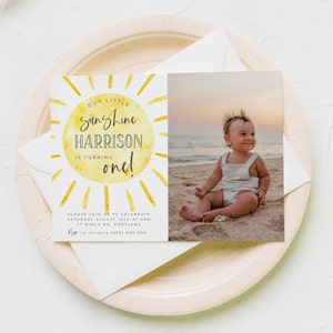 May include: A rectangular birthday invitation card featuring a yellow watercolor sun graphic with the text Our Little Sunshine Harrison Is Turning One. The right side displays a photograph of a child sitting on a beach. Event details include Saturday August 10th at 2pm, 77 Birch Rd, Portland, and an RSVP contact number.