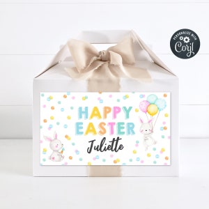 Puede incluir: Caja de regalo blanca con una cinta beige y una etiqueta colorida que dice "Feliz Pascua Juliette" con un conejito sosteniendo una zanahoria y un conejito sosteniendo globos.