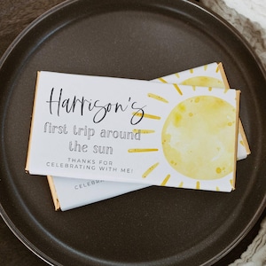 May include: Two white chocolate bar wrappers with gold foil edges are stacked on a dark brown plate. The wrappers feature the text "Harrison's first trip around the sun" and a yellow sun illustration. The text "THANKS FOR CELEBRATING WITH ME!" is also visible.