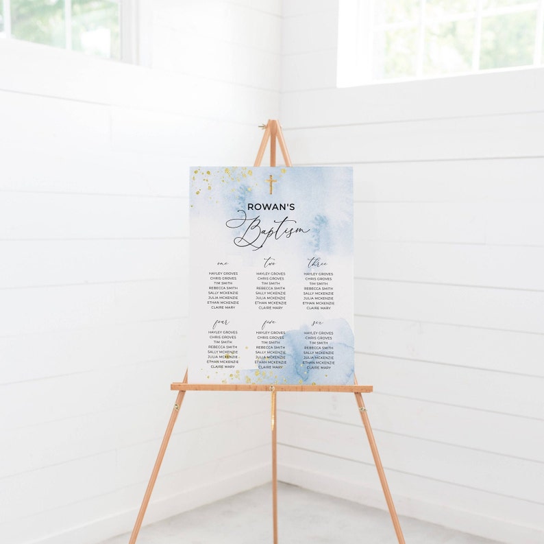 May include: A baptism seating chart on a wooden easel. The sign has a watercolor background in shades of blue and gold. The text "Rowan's Baptism" is displayed at the top, with guest names organized in columns below. A gold cross is at the top.