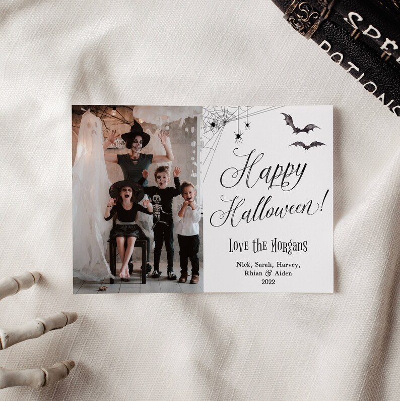 May include: A rectangular Halloween greeting card featuring a photograph of a family in costumes on the left and text on the right. The text reads Happy Halloween! Love the Morgans, Nick, Sarah, Harvey, Rhian & Aiden 2022, with spiderweb and bat illustrations.
