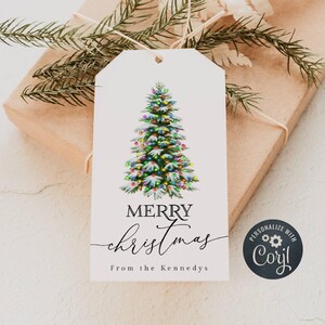 Puede incluir: Una etiqueta de regalo blanca con una ilustración de acuarela de un árbol de Navidad decorado. La etiqueta dice "MERRY Christmas" en cursiva, con "From the Kennedys" debajo. La etiqueta está atada a un regalo envuelto con hilo y ramitas de hoja perenne.