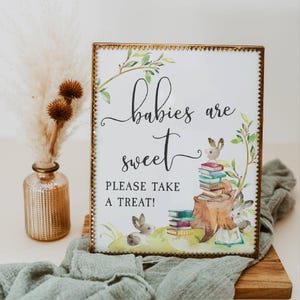 May include: A white sign with a gold frame and a watercolour illustration of bunnies and books. The sign reads "Babies are sweet. Please take a treat!"