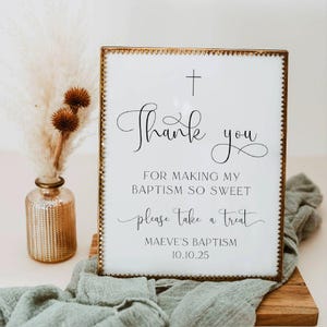 May include: A rectangular sign with a gold metal frame featuring a cross and the black text: Thank you FOR MAKING MY BAPTISM SO SWEET please take a treat MAEVE'S BAPTISM 10.10.25. The sign is displayed on a wooden base with a sage green fabric.