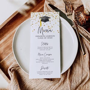 May include: A graduation menu card with a black mortarboard and gold confetti design. The menu lists starters, main courses, and desserts for Annabeth Harrison's Class of 2026. The card is on a white plate with silverware and a decorative cloth.
