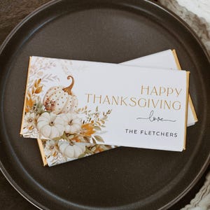 May include: White chocolate bar with a gold foil wrapper featuring a watercolor illustration of pumpkins and fall foliage. The text "Happy Thanksgiving love The Fletchers" is printed in gold on the wrapper.