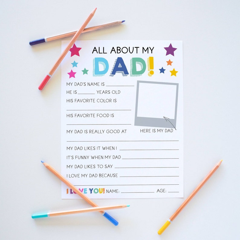 May include: A white paper questionnaire titled ALL ABOUT MY DAD! with colorful stars, blank lines for writing, and a square frame labeled HERE IS MY DAD. The sheet is surrounded by several colored pencils in shades of blue, pink, and yellow.